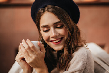 Wall Mural - Joyful young lady with red lips, closed eyes, brown hair, stylish beret and beige pullover sitting outdoors, smiling and holding white cup of tea in sunny autumn day