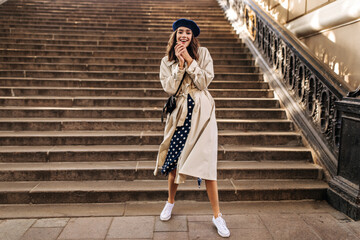 Wall Mural - Cheerful young girl in French beret, beige trench coat, polka dot skirt and white sneakers posing full length, laughing and looking into camera against sunlit stairs background