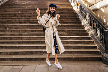Wall Mural - Cute young lady in beige trench, polka dot skirt, beret and white sneakers, smiling, walking around city and posing near sunlit stairs