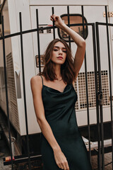 Wall Mural - Gorgeous young brunette with bright makeup, well-built body, silk long slip dress, posing on street, raising one hand and looking into camera