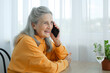 © Ольга  - Beautiful old grandmother with grey hair and face with wrinkles is using smartphone, talking with someone and sitting at the table at home on window background, mother's day, happy retirement