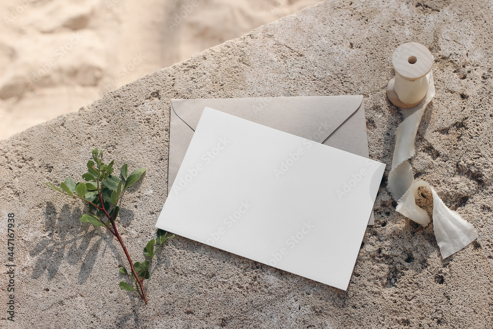 Summer wedding stationery mock-up. Blank greeting card, invitation with craft envelope and silk ribbon. Green lentisk branch on beige concrete background in sunlight. Blurred sand. Flat lay, top view. - obrazy, fototapety, plakaty 