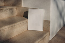 Blank White Picture Frame Leaning Against White Wall. Outdoor Sandstone Stairs In Sunlight, Shadows Overlay. Empty Poster Mockup For Art Display. Minimal Summer Design. No People.