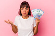 © Asier - Young mixed race woman holding bills isolated on pink background shrugs shoulders and open eyes confused.