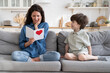 © DimaBerlin - Surprised young mother reading congratulation from gift postcard with heart sitting near little son congratulate happy single mom with mother's day or birthday. Cute child make surprise to mum at home