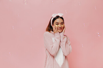 Wall Mural - Young charming Asian woman in kigurumi smiles on pink background. Brunette girl in pajamas and soft headband touches cheeks and poses with cosmetic eyepatches on isolated backdrop with bubbles.