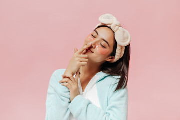 Wall Mural - Brunette pretty woman in blue pajamas and soft beige headband touches nose. Cute Asian girl in sleep wear poses with eyepatches on pink background.