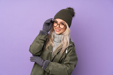 Wall Mural - Teenager blonde girl with winter hat over isolated purple background with glasses and happy