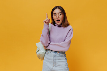 Wall Mural - Teen cool brunette Asian woman in eyeglasses, purple sweater and denim skirt points up, has great new idea and poses with backpack on orange background.