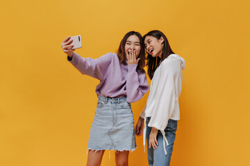 Happy girls in sweatshirts take selfie and laugh on orange background. Young woman in purple sweater and denim skirt holds phone on isolated.