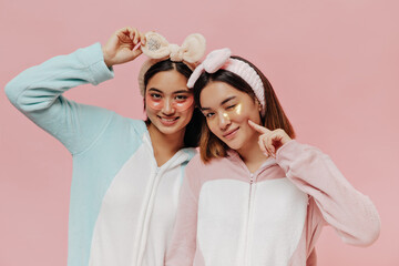 Wall Mural - Pretty brunette Asian girls in pajamas, soft headbands and eyepatches smile and pose on isolated pink background.