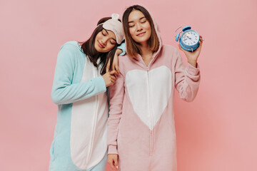 Wall Mural - Friend in pajamas pose with closed eyes on pink background. Short-haired girls in sleep wear hold blue alarm clock on isolated.