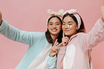 Wall Mural - Pretty happy brunette girls in kigurumis take selfie on pink background. Charming young women in pajamas and headbands smile.