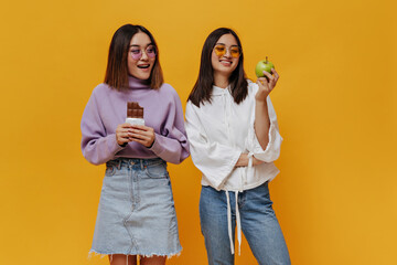 Wall Mural - Charming Asian girl in denim skirt and purple sweater holds milk chocolate bar and looks at fruit of her friend. Brunette woman in jeans and white hoodie hold fresh green apple.