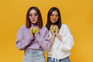 Wall Mural - Portrait of brunette young Asian girl in colorful sunglasses and sweatshirts hold green fresh apple on isolated orange background.