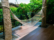 Empty white cotton rope cradle. Swing hammock hanging between two palm trees at the wooden terrace near outdoor garden.