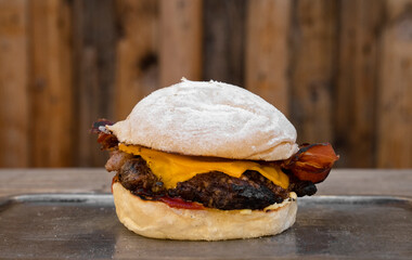 American food. Closeup view of a cheeseburger with cheddar cheese, meat, bread, crispy bacon and sauce, in a metal dish on the table with a wooden background.  