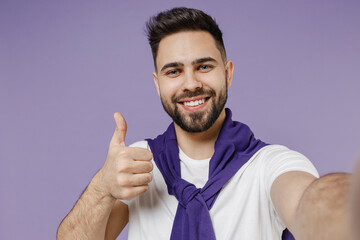 Wall Mural - Close up beautiful smiling young brunet man 20s wears white t-shirt purple shirt doing selfie shot on mobile phone showing thumb up like gesture isolated on pastel violet background studio portrait