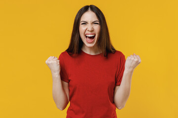 Beautiful fun young brunette woman 20s wears basic red t-shirt do winner gesture celebrating clenching fists say yes isolated on yellow background studio portrait. People emotions lifestyle concept