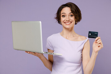 Poster - Young smiling happy woman 20s wear white t-shirt using laptop pc computer hold credit bank card doing online shopping order delivery booking tour isolated on pastel purple background studio portrait