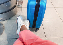 An Unrecognizable Traveler In Pink Trousers And White Sneakers, Sitting In A Relaxed Pose, With Her Feet Placed Next To A Blue Suitcase, Waiting For A Trip