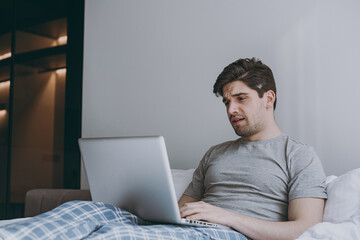 Sad disappointed excited young man 20s wearing pajamas grey t-shirt sit in bed hold use work on laptop pc computer rest relax at home indoors bedroom. Good mood sleeping night morning bedtime concept