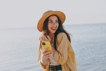 Young smiling happy surprise woman in straw hat shirt summer casual clothes using mobile cell phone browsing outdoors at sunrise sun dawn over sea background People vacation lifestyle journey concept