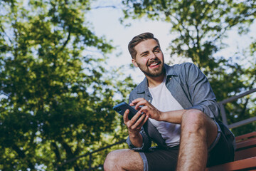 Wall Mural - Bottom view young smiling minded fun man 20s in blue shirt shorts sit on bench use mobile cell phone look aside rest relax in spring green city park outdoor on nature Urban leisure lifestyle concept.