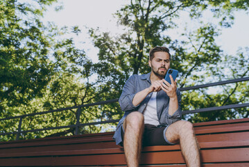 Wall Mural - Bottom view young serious man 20s in blue shirt shorts sit on bench using mobile cell phone chat online browsing rest relax in spring green city park outdoor on nature Urban leisure lifestyle concept.