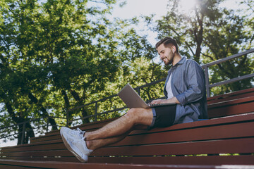 Wall Mural - Bottom view full body young fun man in blue shirt shorts earphones sit on bench work online laptop pc computer waving hand rest relax in spring green city park outdoor on nature Urban leisure concept.