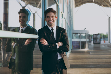 Smiling cheerful young traveler brunet businessman man 20s wear black classic tie suit standing outside at international airport terminal look aside. People air flight business trip lifestyle concept.