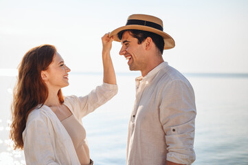 Side view smiling happy young couple two family man woman in casual clothes girlfriend put hat on boyfriend rest date at sunrise over sea sand beach ocean outdoor seaside in summer day sunset evening.