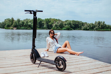 A beautiful brunette girl rests on a wooden bridge near the river next to an electric scooter. The concept of ecology and electric transport