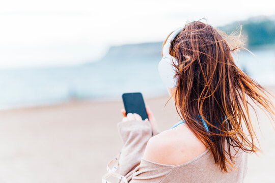 beautiful young adult woman listening to music on the beach on a cloudy summer or spring day wearing headphones