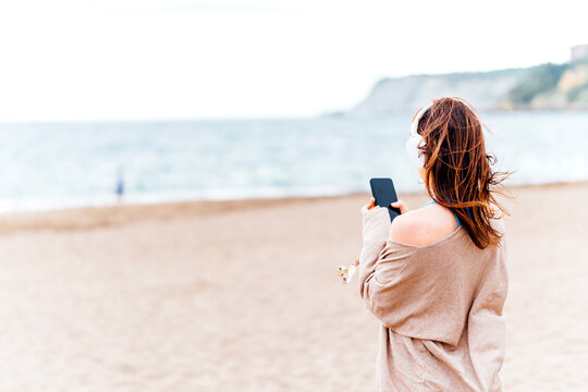 beautiful young adult woman listening to music on the beach on a cloudy summer or spring day wearing headphones
