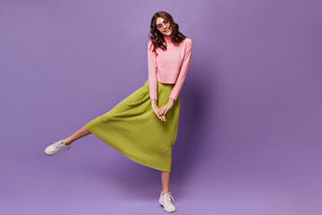 Wall Mural - Pretty curly woman rises leg. Attractive young girl in green midi skirt and pink sweater smiles and moves on purple background.
