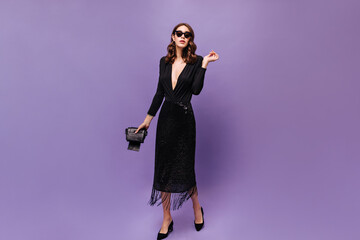 Wall Mural - Beautiful curly lady in festive dress moves on purple background. Stylish woman in shiny skirt and black blouse poses in sunglasses.