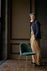 Full length shot of fashionable grey haired mature man in casual wear looking out a window while standing in modern loft interior