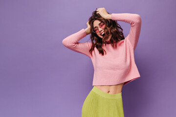 Wall Mural - Excited brunette woman ruffles hair on purple background. Cheerful girl in pink sweater and green skirt laughs on isolated.