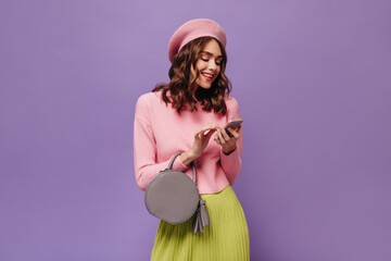Wall Mural - Charming curly woman in green skirt, pink beret and sweater chats and holds phone. Beautiful lady poses with handbag on purple background.
