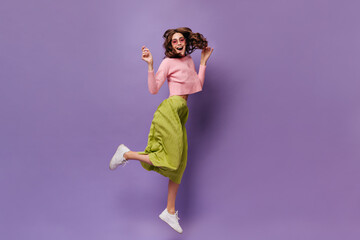 Wall Mural - Joyful brunette woman in green skirt and pink sweater jumps on purple background. Happy curly girl smiles and moves on isolated.