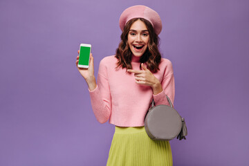 Wall Mural - Excited brunette woman in green skirt and pink sweater points at phone screen. Charming girl in beret holds grey handbag and smiles on purple background.