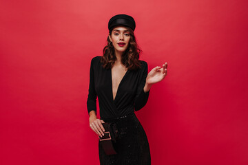 Happy lady with bright lips looks into camera on red background. Elegant woman in black dress and cap poses on isolated.