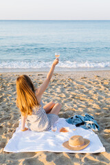 Wall Mural - Summer picnic on the beach at sunset. Young woman with glass of rose wine. Weekend picnic. Rear view, people from behind.