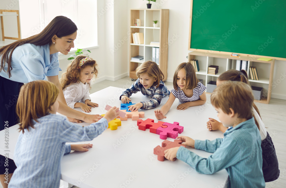 Team of creative little children with teacher join and fit together big colorful jigsaw puzzle standing around white table in classroom at school or educational center. Education and teamwork concept - obrazy, fototapety, plakaty 