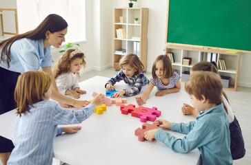 Team of creative little children with teacher join and fit together big colorful jigsaw puzzle standing around white table in classroom at school or educational center. Education and teamwork concept