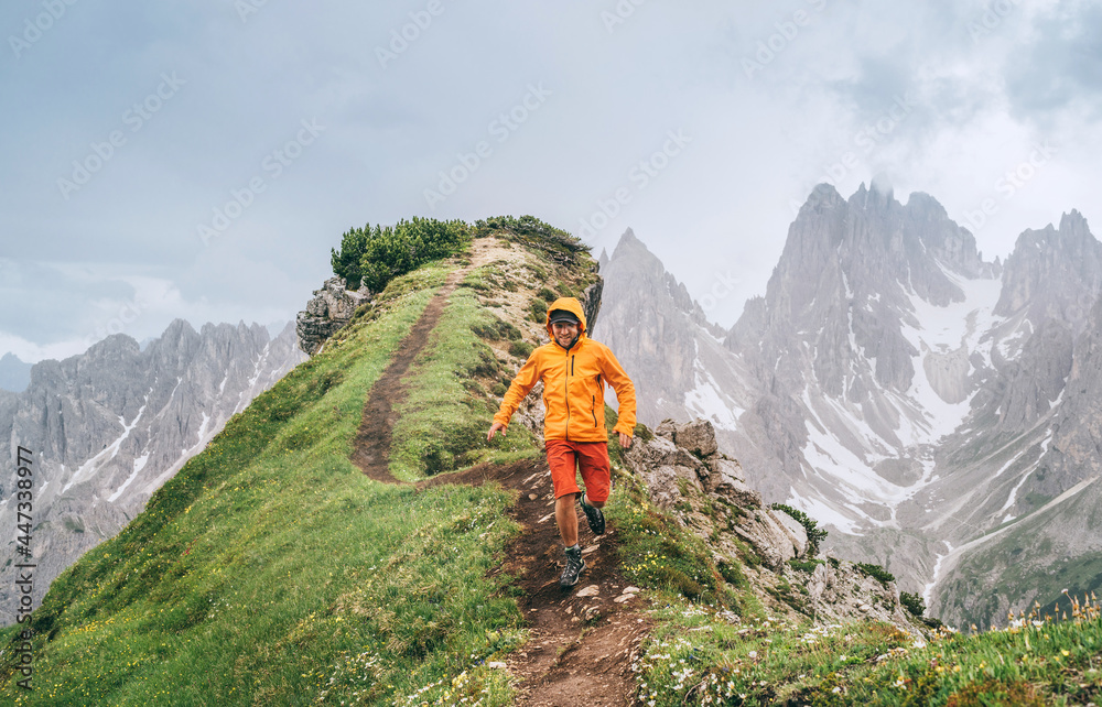 Dressed bright orange soft shell jacket backpacker running  by green mountain path with picturesque Dolomite Alps range background,. Active people and European mountain hiking tourism concept image. - obrazy, fototapety, plakaty 