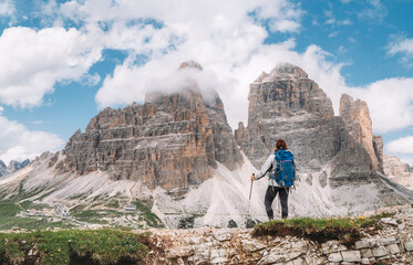 Wall Mural - Female hiker walking with backpack and trekking poles on green mountain hill enjoying picturesque Tre Cime di Lavaredo formation 2999m in Dolomite Alps. Active people and mountain concept