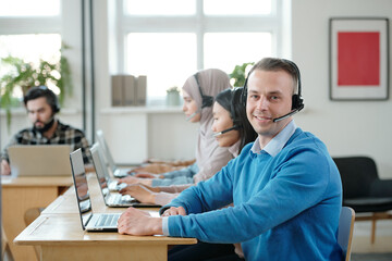 Poster - Young successful operator with headset looking at you with smile against his co-workers