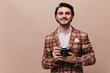 Gallant young brunette-haired gentleman in plaid blazer and stylish shirt looking straight and holding camera. Man posing isolated over beige background